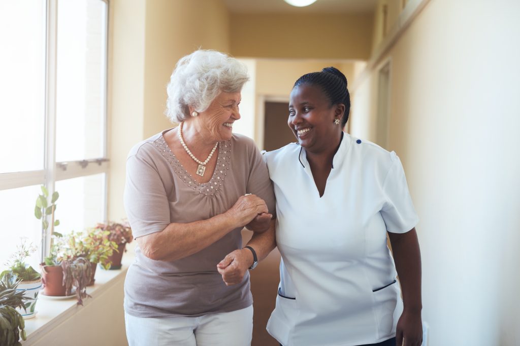Portrait of smiling senior living caregiver and senior woman walking together through a corridor of a retirement home. Healthcare worker taking care of elderly woman.