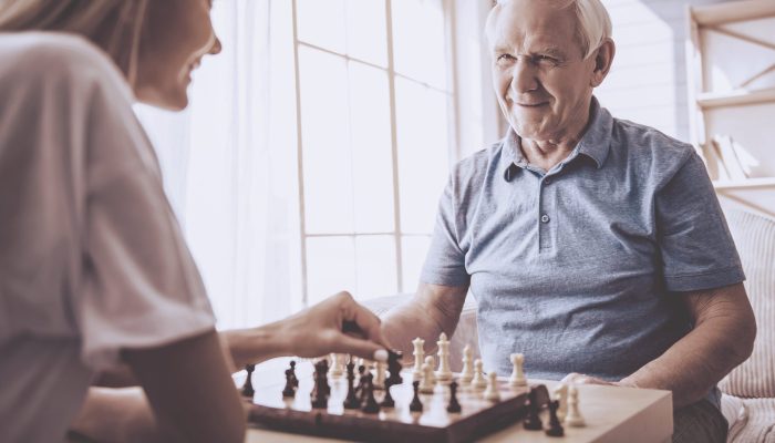Beautiful young girl-volunteer and handsome old man are playing chess and smiling